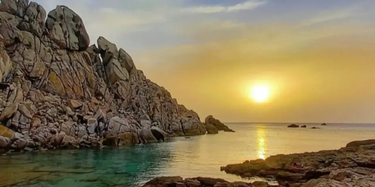 Scogliere rocciose bagnate da acque cristalline, con il sole che tramonta sulla valle della luna, creando riflessi dorati sul mare.