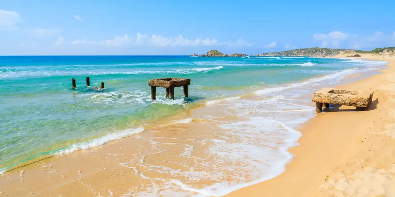 Spiaggia di Chia, Cagliari. Resti di antiche strutture in mare e sulla sabbia dorata, con acque turchesi e cielo azzurro.