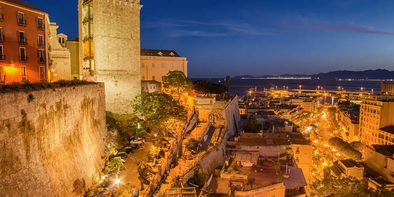 Torre medievale e palazzi illuminati nella piazza di Cagliari, con il porto sullo sfondo al crepuscolo.