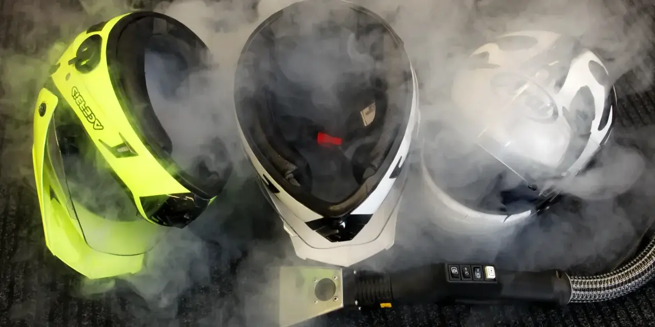 Tre caschi, uno giallo fluo, uno bianco e nero, e uno bianco, sono avvolti dal vapore. Un accessorio per la pulizia a vapore è posizionato davanti a loro, suggerendo che il casco non deve essere pulito con solventi.