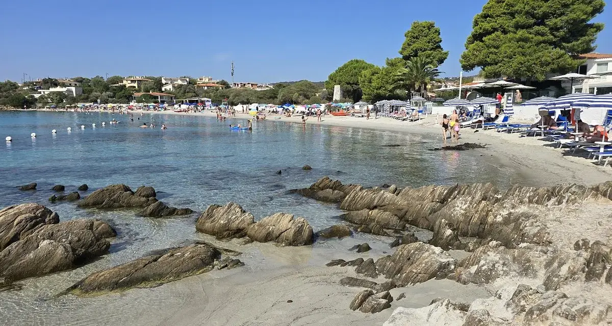 Spiaggia del Pellicano: sabbia chiara, rocce levigate e mare cristallino con bagnanti e ombrelloni.