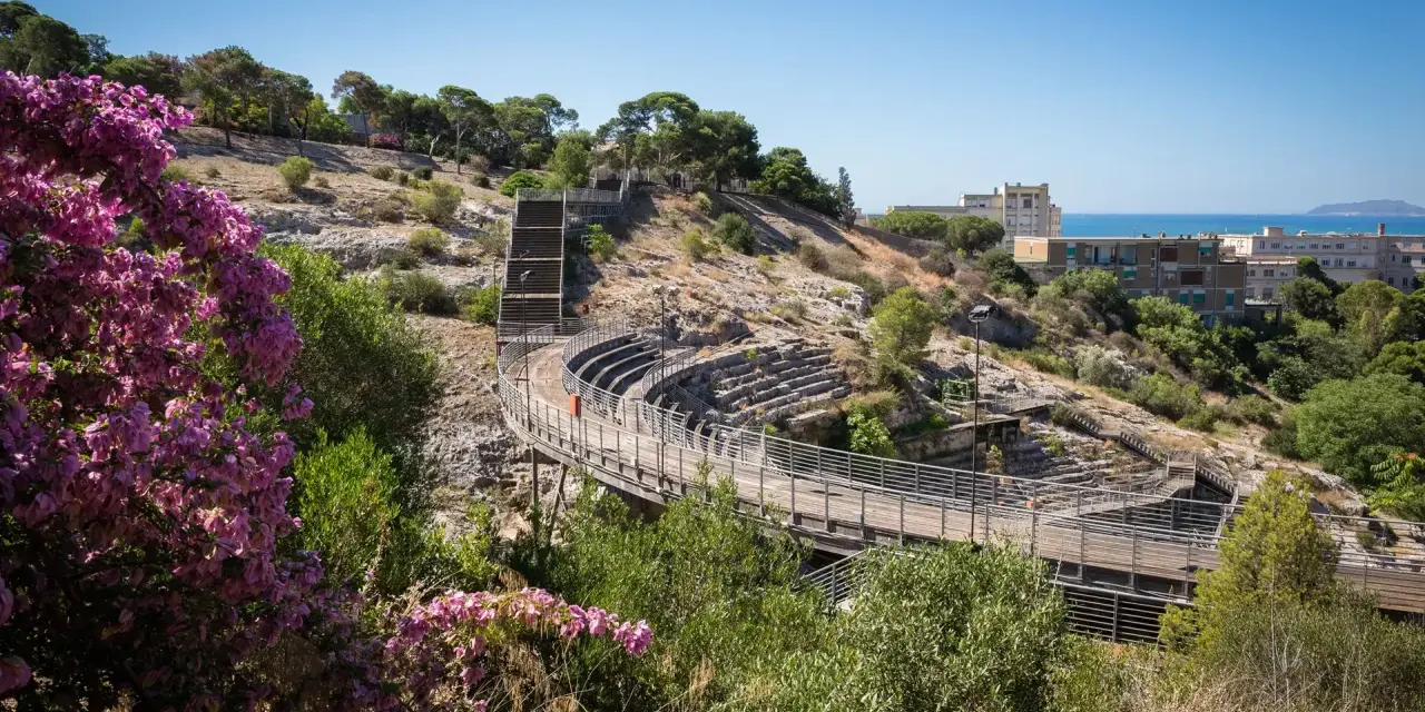 Un suggestivo anfiteatro a Cagliari, con gradinate in pietra e passerelle in legno, immerso nel verde e con fiori viola in primo piano.