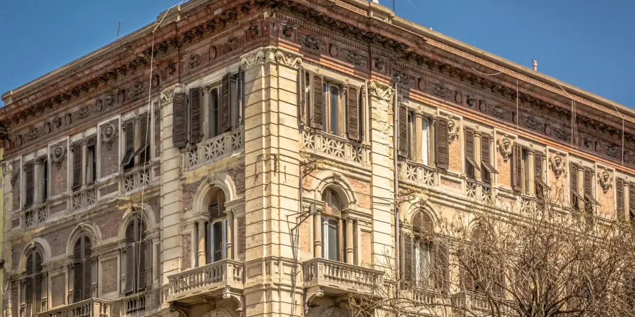 Angolo del **Palazzo Accardo a Cagliari**, con facciata in pietra chiara, balconi in ferro battuto e finestre con persiane.