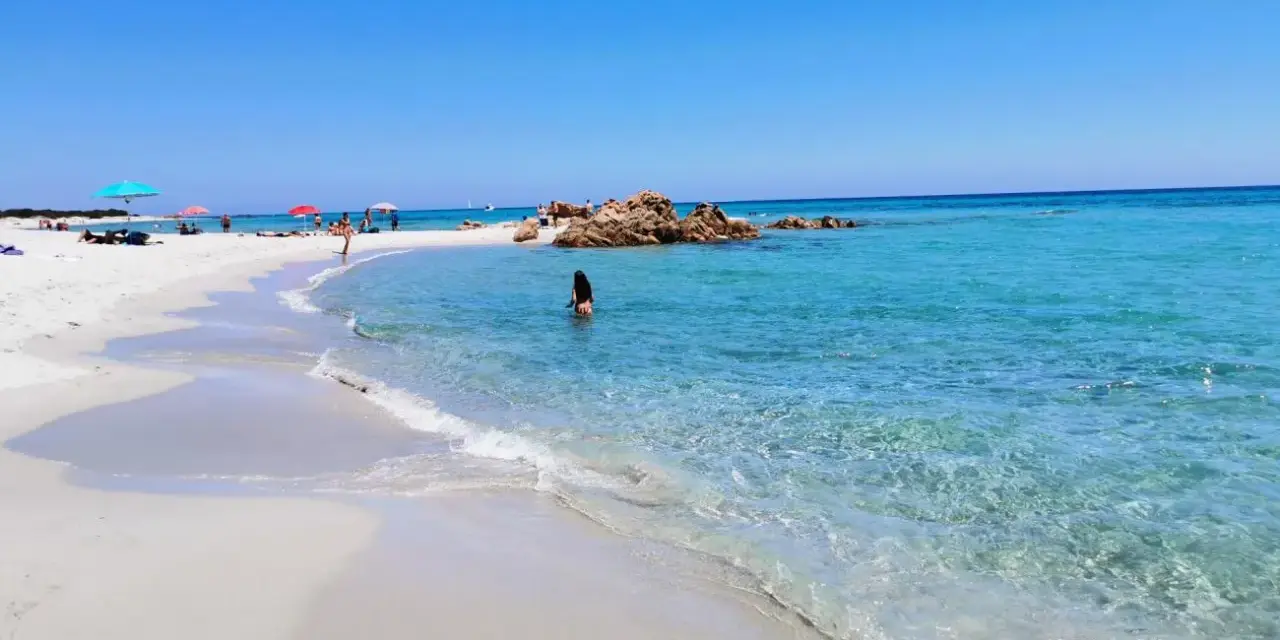Spiaggia di Berchida: sabbia bianca, acque cristalline e rocce rosse sotto un cielo azzurro. Una donna si gode il mare calmo.