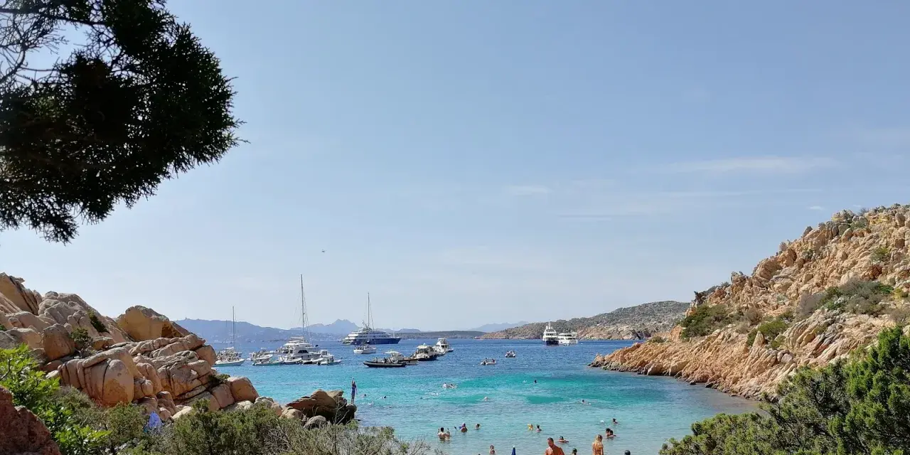 Spiaggia affollata a Cala Coticcio, con gente che prende il sole e nuota nelle acque cristalline. Diverse barche e yacht ancorati nella baia.