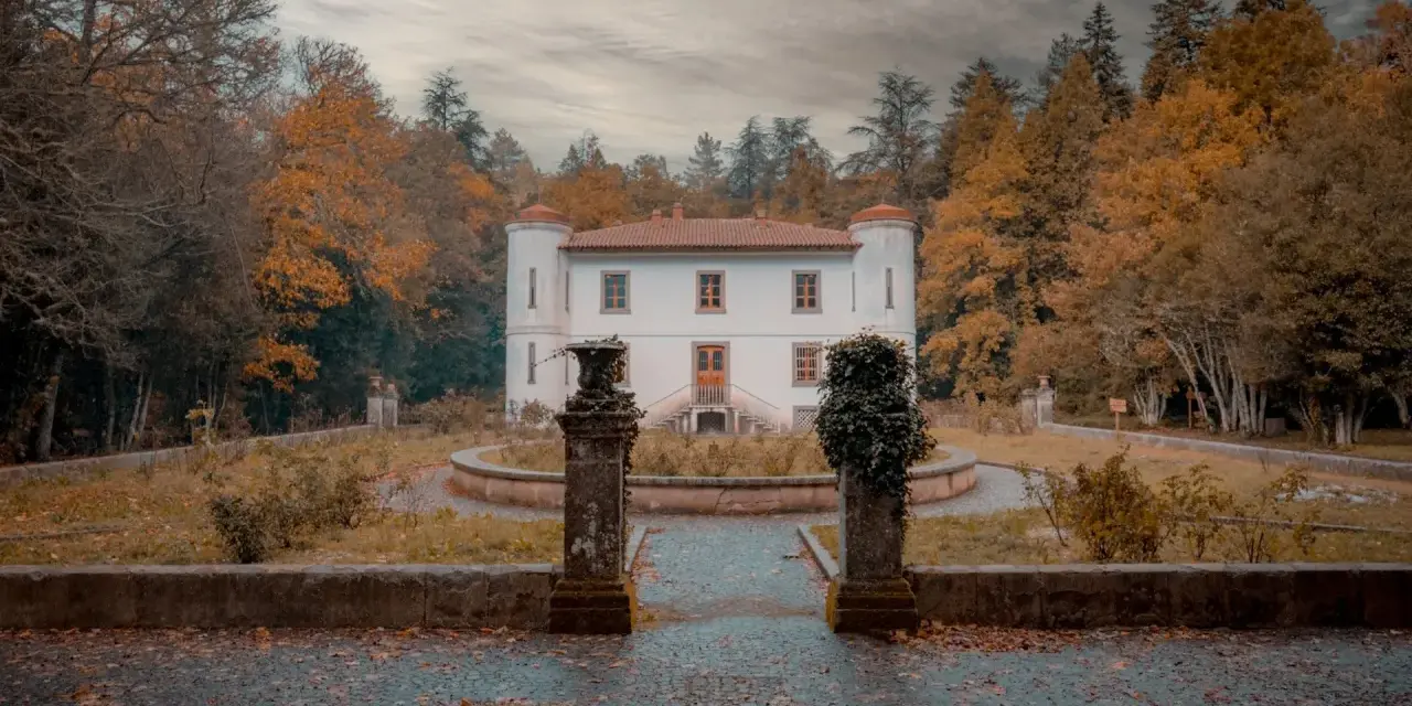 Un'antica villa bianca con torrette, circondata da alberi autunnali dai colori caldi. Un sentiero lastricato conduce all'ingresso, tra statue e vegetazione. Le **badde salighes** sussurrano storie di tempi passati.