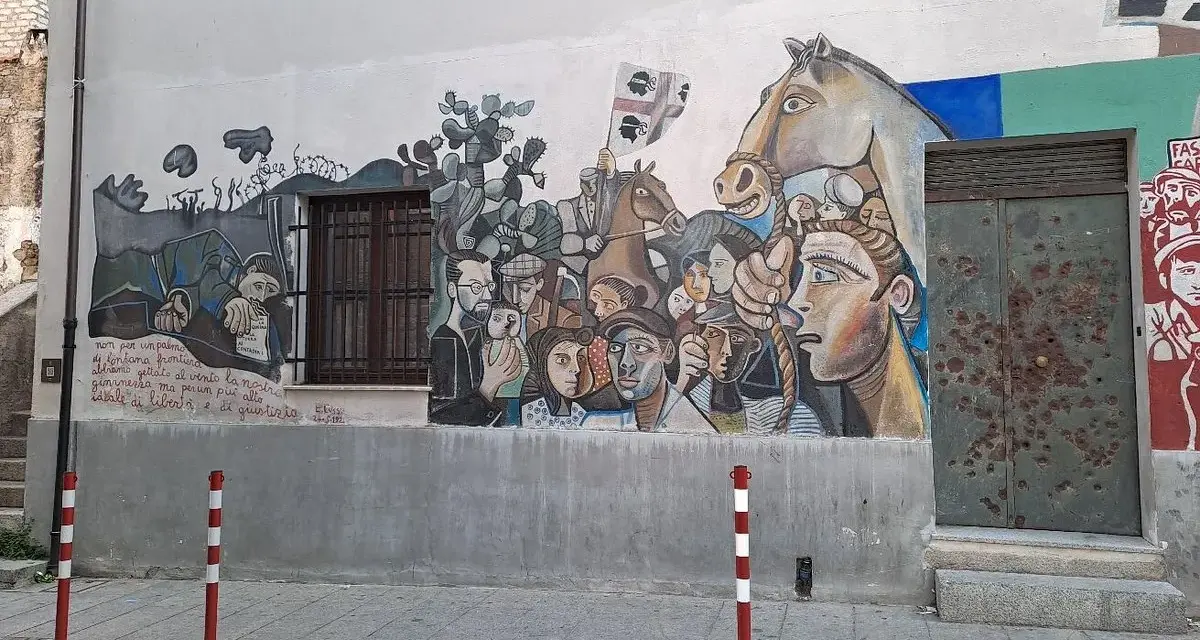 Un grande murale colorato decora un edificio in un paese, con volti, un cavallo e bandiere. Un'opera d'arte che racconta storie di un paese, murales in Sardegna.