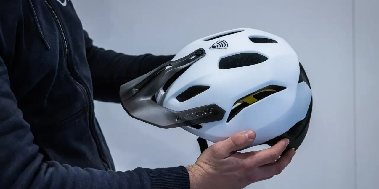 Mani che tengono la calotta esterna di un casco da bici bianco con visiera scura e logo Dainese.