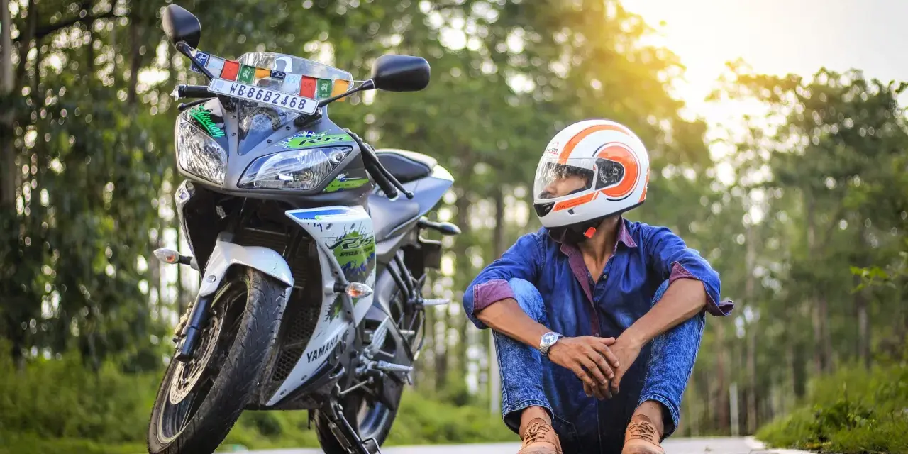 Giovane con casco da moto arancione e bianco, seduto accanto alla sua Yamaha. Spera di allargare il casco stretto per un viaggio più comodo.