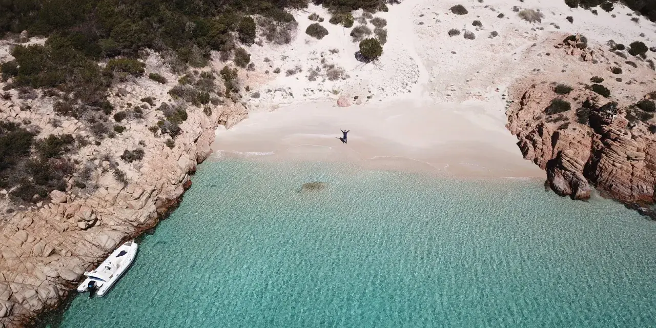 Una persona celebra la bellezza di Cala Soraya, con acque cristalline e rocce rosse. Una barca è ormeggiata.