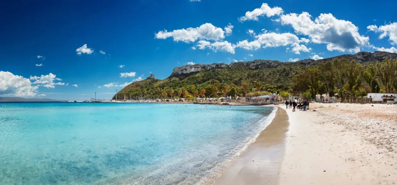 Spiaggia con acque cristalline e la sagoma della Sella del Diavolo all'orizzonte, sotto un cielo azzurro con nuvole bianche.