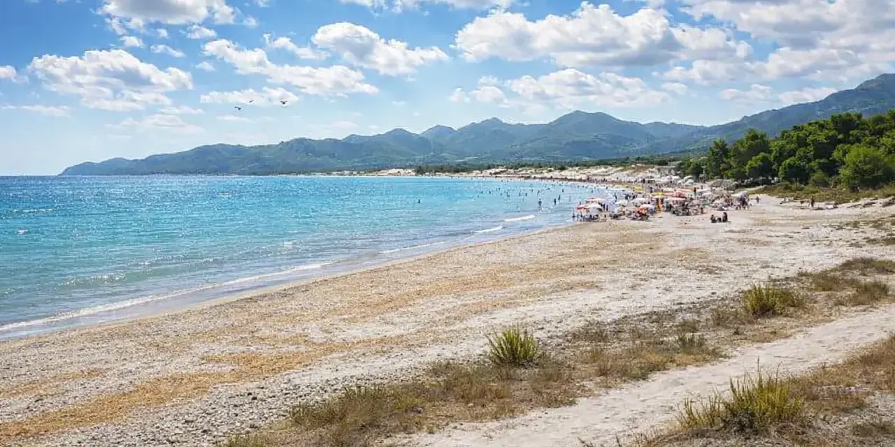 Spiaggia affollata con ombrelloni e gente che si gode il sole. Il nuoro mare azzurro lambisce la costa, con montagne verdi sullo sfondo.