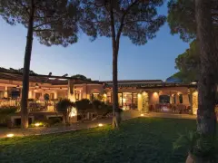 La sorgente Villasimius: un ristorante con tavoli all'aperto illuminati da luci soffuse, circondato da pini e un prato curato.
