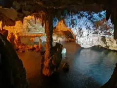 Interno di una grotta con stalattiti e stalagmiti illuminate da luci arancioni e blu. L'acqua scura riflette le formazioni rocciose in questa grotta verde.