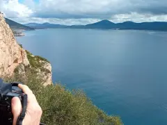 Scogliera a picco sul mare blu, con un fotografo che cattura il paesaggio. Si intravede la costa e le montagne all'orizzonte, forse vicino alla grotta verde di Alghero.