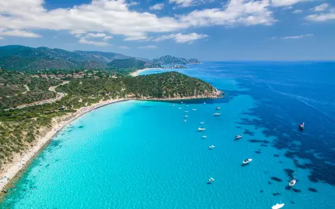 Spiaggia di Chia, Sardegna: acque cristalline che sembrano mari pintau, con barche a vela e un litorale affollato.