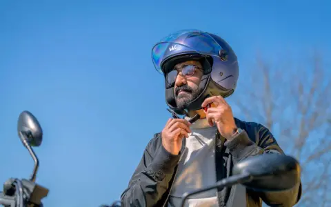 Uomo con barba e occhiali da sole che regola il cinturino di un casco da moto blu iridescente. La corretta misura casco moto è fondamentale per la sicurezza.