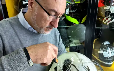 Uomo con occhiali regola un casco da moto bianco, mentre altri caschi sono esposti. Si nota l'anno produzione casco moto su un'etichetta.