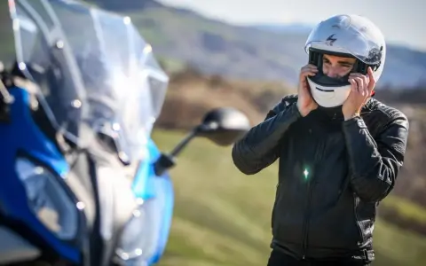 Uomo in giacca di pelle che indossa caschi per moto bianchi, pronto per un viaggio su una moto blu sfocata.