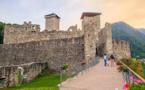 Un sentiero fiorito conduce al maestoso Castello San Michele, con le sue torri imponenti e mura antiche che si stagliano contro il cielo.