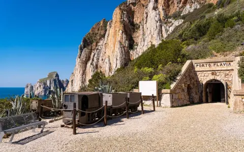 Antichi vagoni minerari e l'ingresso di Porto Flavia, testimoni della storica zona mineraria sarda, si stagliano contro un imponente promontorio roccioso.
