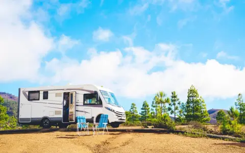 Un camper bianco è parcheggiato in un'area sterrata con due sedie blu e un tavolino. Sullo sfondo, pini e colline sotto un cielo azzurro con nuvole bianche. Un luogo ideale per un soggiorno in stile piscin'e janas.