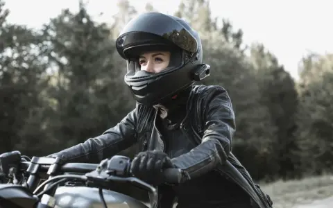 Woman in black leather jacket and helmet riding a motorcycle outdoors.