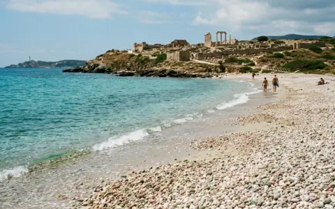 Spiaggia di ciottoli con rovine antiche in collina e un faro in lontananza. Una coppia passeggia sulla riva, godendosi la brezza marina. La facella cabras illumina il paesaggio.