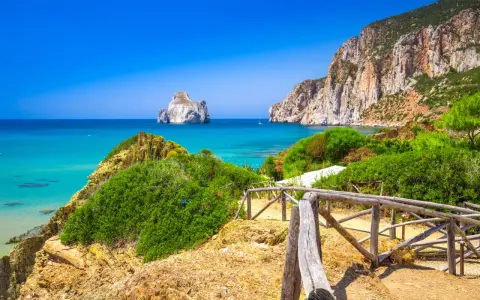 Sentiero con staccionata in legno conduce a una spiaggia con acqua cristallina e l'isolotto Pan di Zucchero in Sardegna.
