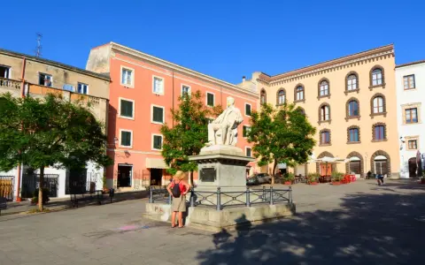 La piazza Tola a Sassari, con il monumento al centro, edifici colorati e alberi verdi sotto un cielo azzurro.