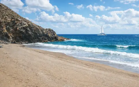 Spiaggia dorata e mare azzurro vicino a Sassari, con un catamarano bianco che naviga all'orizzonte sotto un cielo nuvoloso.