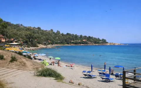 Spiaggia di Porto Frailis con ombrelloni colorati, lettini blu e gente che si gode il mare cristallino sotto il sole.
