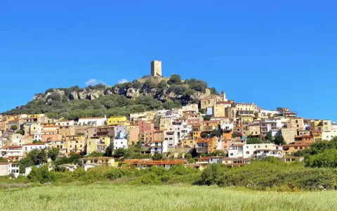 La **posada di sera** si staglia su una collina rocciosa, con case colorate e una torre medievale sotto un cielo azzurro.