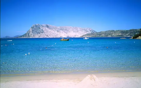 Spiaggia di Cala Suaraccia, mare cristallino, montagne sullo sfondo e barche in lontananza. Come arrivare: goditi il panorama!