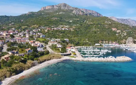 Vista aerea del centro storico di Santa Maria Navarrese, con case arroccate sulla collina, un porto turistico pieno di barche e il mare cristallino.
