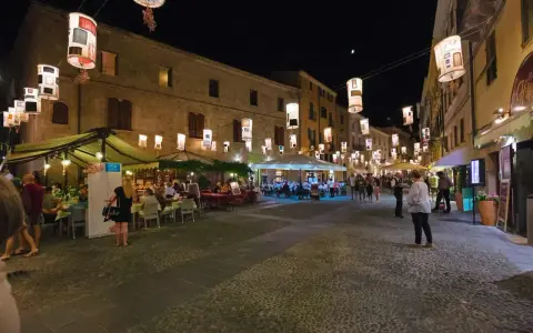 Serata ad Alghero: ristoranti all'aperto illuminati da lanterne, gente che passeggia e cena.