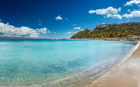 Acque cristalline e sabbia dorata sulle spiagge di Cagliari, con il promontorio verdeggiante e il porto turistico sullo sfondo.
