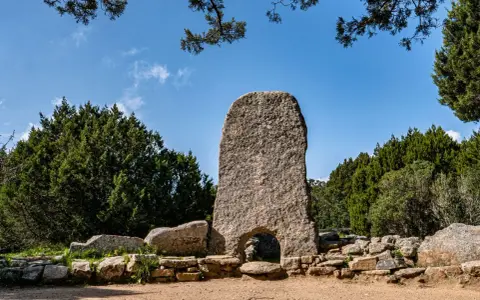 La tomba dei giganti in Sardegna, un imponente monolito di granito con un arco, circondato da pietre antiche e vegetazione rigogliosa sotto un cielo azzurro.