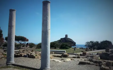 Colonne antiche nel **sito archeologico** di Nora, con una torre in lontananza su un'isola rocciosa.