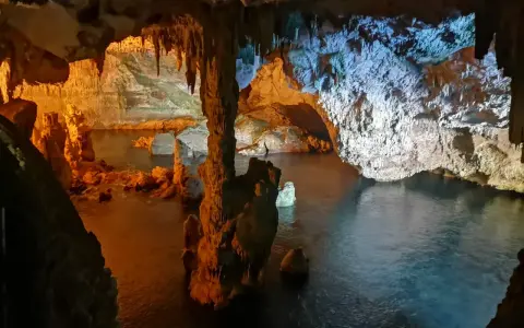 Interno di una grotta con stalattiti e stalagmiti illuminate da luci arancioni e blu. L'acqua scura riflette le formazioni rocciose in questa grotta verde.