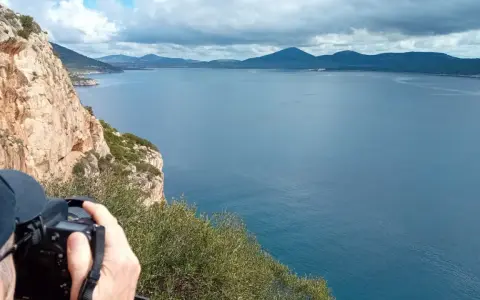 Scogliera a picco sul mare blu, con un fotografo che cattura il paesaggio. Si intravede la costa e le montagne all'orizzonte, forse vicino alla grotta verde di Alghero.