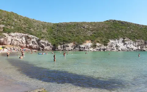 Persone si godono il sole e il mare cristallino nella spiaggia cala lunga, con colline verdi e rocce a fare da cornice.