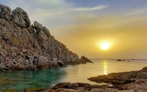 Scogliere rocciose bagnate da acque cristalline, con il sole che tramonta sulla valle della luna, creando riflessi dorati sul mare.