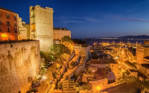 Torre medievale e palazzi illuminati nella piazza di Cagliari, con il porto sullo sfondo al crepuscolo.