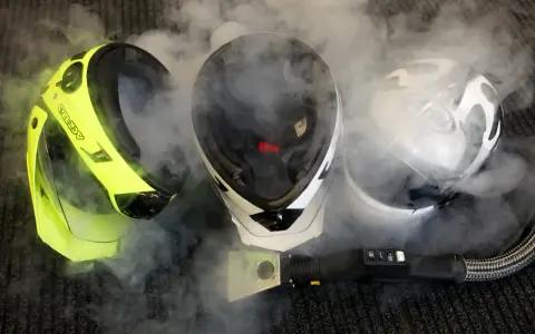 Tre caschi, uno giallo fluo, uno bianco e nero, e uno bianco, sono avvolti dal vapore. Un accessorio per la pulizia a vapore è posizionato davanti a loro, suggerendo che il casco non deve essere pulito con solventi.
