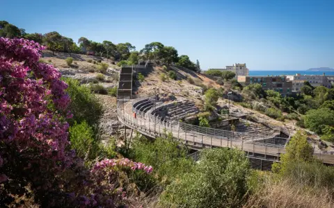 Un suggestivo anfiteatro a Cagliari, con gradinate in pietra e passerelle in legno, immerso nel verde e con fiori viola in primo piano.