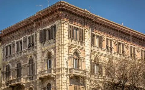 Angolo del **Palazzo Accardo a Cagliari**, con facciata in pietra chiara, balconi in ferro battuto e finestre con persiane.