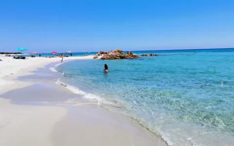 Spiaggia di Berchida: sabbia bianca, acque cristalline e rocce rosse sotto un cielo azzurro. Una donna si gode il mare calmo.