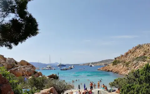 Spiaggia affollata a Cala Coticcio, con gente che prende il sole e nuota nelle acque cristalline. Diverse barche e yacht ancorati nella baia.