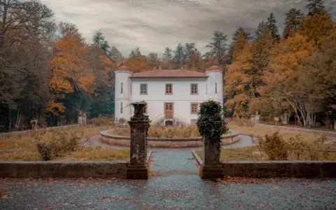 Un'antica villa bianca con torrette, circondata da alberi autunnali dai colori caldi. Un sentiero lastricato conduce all'ingresso, tra statue e vegetazione. Le **badde salighes** sussurrano storie di tempi passati.